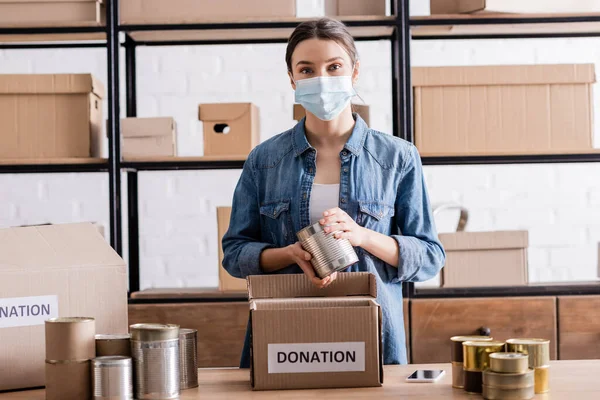 Venditore in maschera medica che tiene cibo in scatola vicino alla scatola con lettere di donazione nel negozio online — Foto stock