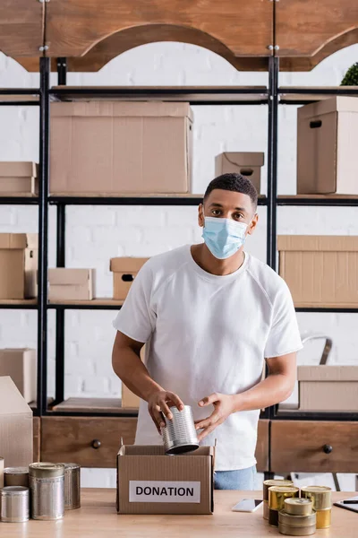 African american seller in medical mask holding can near box with donation lettering in online web store — Stock Photo