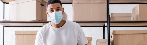 Afrikanisch-amerikanischer Verkäufer mit medizinischer Maske schaut in die Kamera in der Nähe von Schachteln auf dem Regal im Online-Webshop, Banner — Stockfoto
