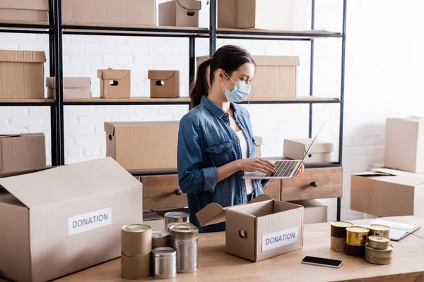 Joven vendedor en máscara médica utilizando el ordenador portátil cerca de cajas con letras de donación y alimentos enlatados en la tienda web en línea - foto de stock