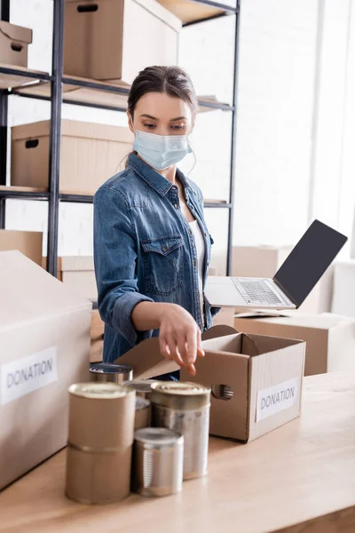 Seller in medical mask pointing at canned food near donation boxes and holding laptop in online web store — Stock Photo