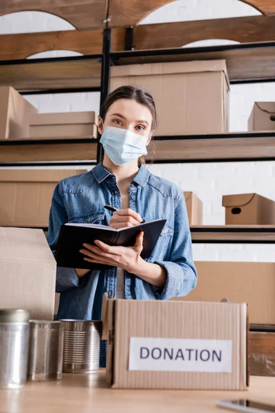 Joven vendedor en la escritura de máscara médica en cuaderno cerca de caja con letras de donación y alimentos enlatados en la tienda en línea web - foto de stock