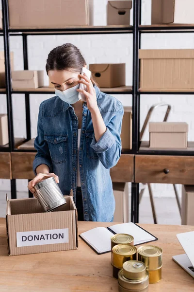 Seller in medical mask talking on smartphone and holding canned food near box with donation lettering on table — Stock Photo
