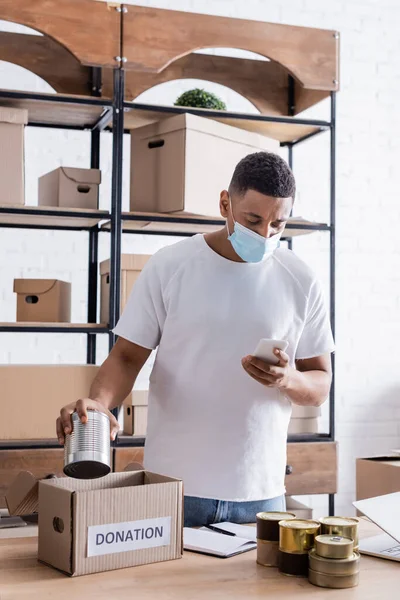 Venditore afroamericano in maschera medica con smartphone e cibo in scatola vicino alla scatola delle donazioni sul tavolo — Foto stock