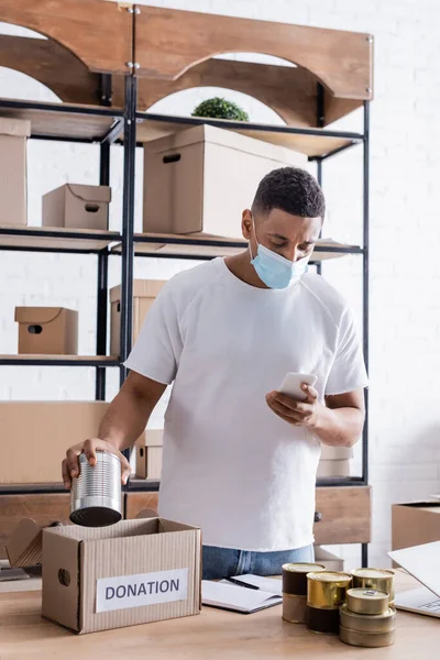 Afrikanischer Verkäufer in medizinischer Maske hält Smartphone und Konserven in der Nähe der Spendenbox auf dem Tisch — Stockfoto