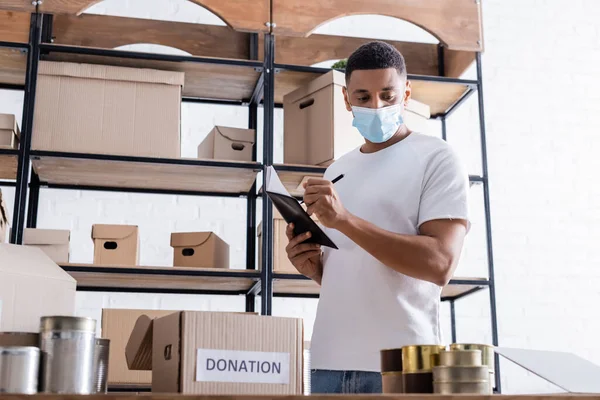 African american online web store owner writing on notebook near canned food and donation box — Stock Photo