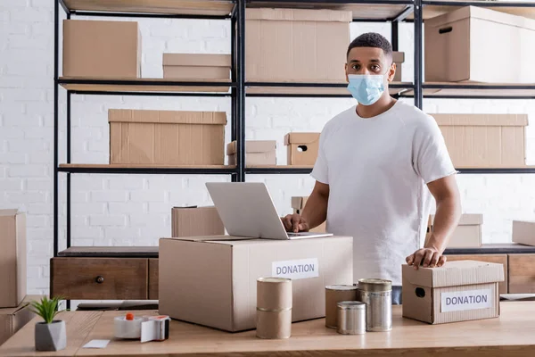 African american owner in medical mask looking at camera near laptop, cans and donation boxes in online web store — Stock Photo