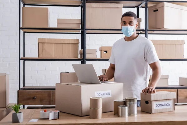 Propietario afroamericano en máscara médica mirando la cámara cerca de la computadora portátil, latas y cajas de donación en la tienda web en línea - foto de stock