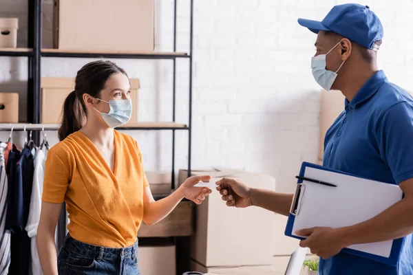 Seller in medical mask taking business card from african american courier in online web store — Stock Photo