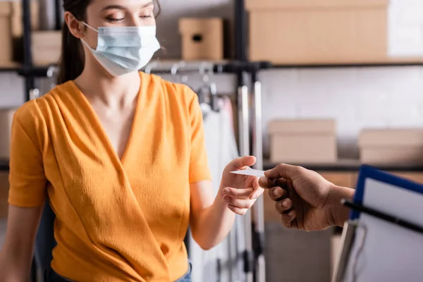 Mensajero afroamericano dando tarjeta de visita al vendedor en máscara médica en la tienda web en línea - foto de stock