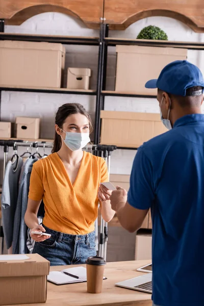 Seller in medical mask holding smartphone and business card near african american courier in online web store — Stock Photo