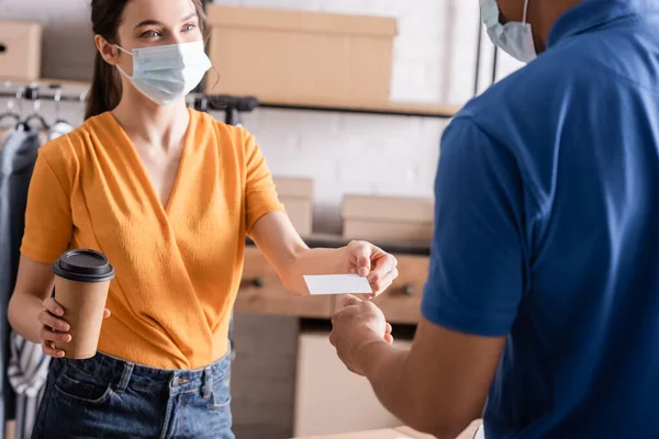 Venditore in maschera medica in possesso di caffè per andare vicino corriere americano africano nel negozio online web — Foto stock