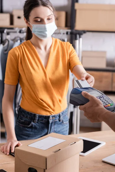 Seller in medical mask paying with credit card near african american courier and carton box in online web store — Stock Photo