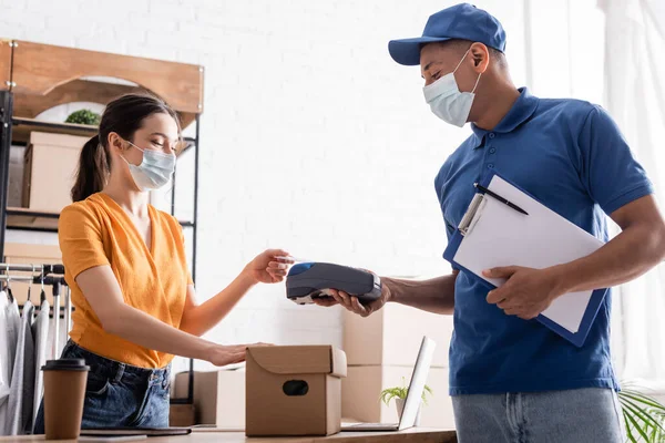 Vendedor en máscara médica pagando con tarjeta de crédito cerca de mensajero afroamericano y caja de cartón en tienda web en línea - foto de stock
