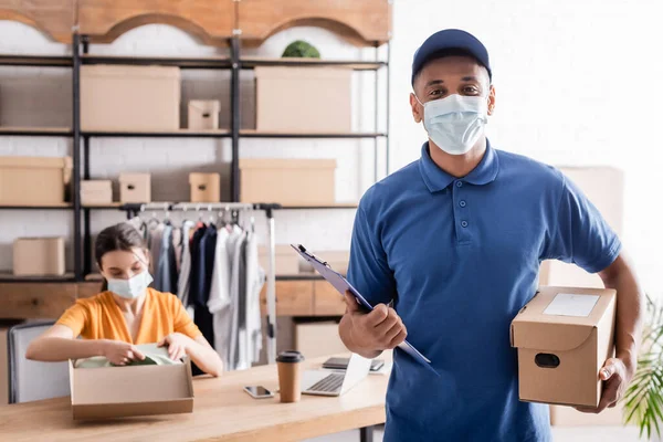 Courrier afro-américain en masque médical tenant presse-papiers et boîte près du vendeur flou dans la boutique en ligne — Photo de stock