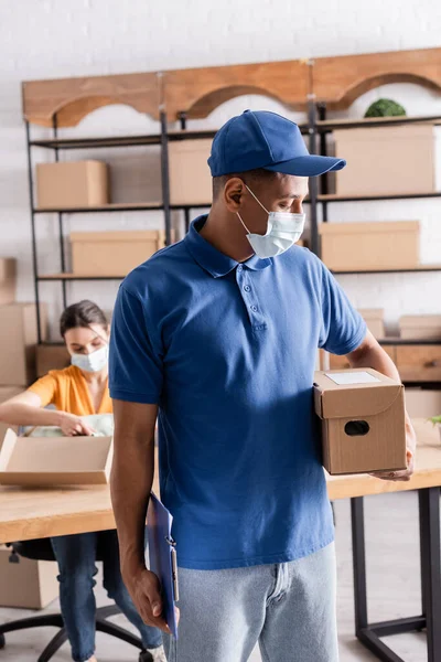 Mensajero afroamericano en máscara médica que sostiene la caja de cartón cerca del dueño borroso de la tienda web en línea - foto de stock