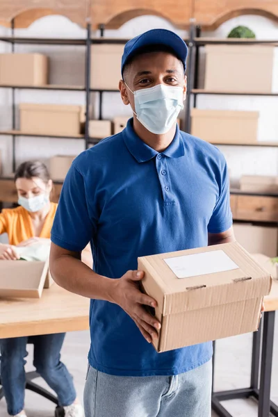 African american courier in medical mask holding cardboard box in online web store — Stock Photo