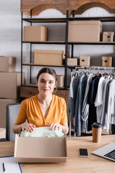 Propietario sonriente poniendo ropa en la caja cerca de dispositivos y café para ir en la tienda web en línea - foto de stock