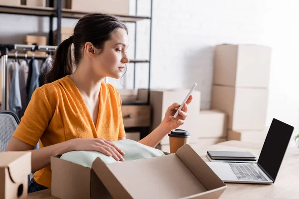 Vista lateral del propietario usando teléfono inteligente cerca de la ropa en caja de cartón en la tienda web en línea - foto de stock