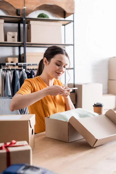 Propietario sonriente tomando fotos en ropa en caja de cartón en tienda web en línea - foto de stock