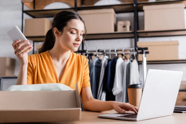 Propietario utilizando dispositivos cerca de café para ir y caja de cartón en la tienda web en línea - foto de stock