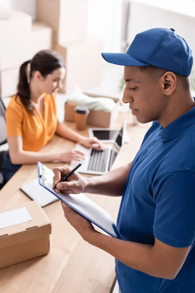 Africano americano entregador escrevendo na área de transferência na loja online — Fotografia de Stock