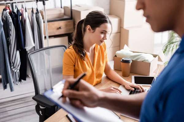 Inhaber benutzt Laptop in der Nähe von verschwommenem afrikanisch-amerikanischem Kurier mit Klemmbrett in Online-Webshop — Stockfoto