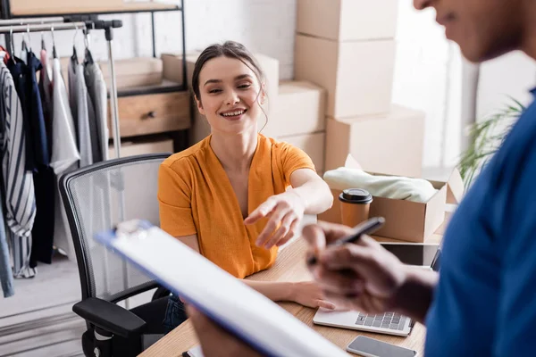 Lächelnder Inhaber zeigt auf Klemmbrett in der Nähe verschwommener afrikanisch-amerikanischer Kurier in Online-Webshop — Stockfoto