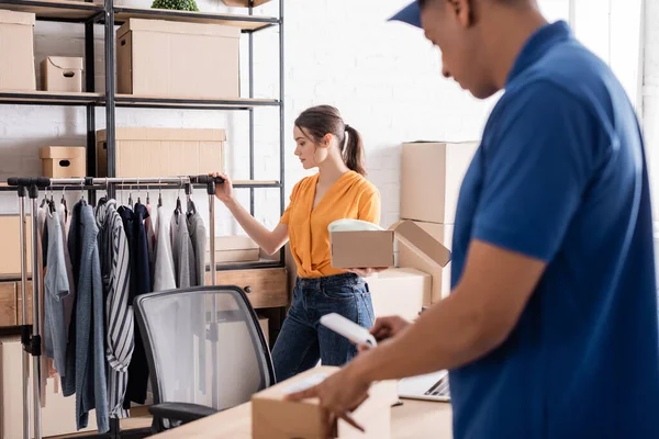 Verkäufer steht neben Kleidung und verschwommenem afrikanisch-amerikanischem Kurier mit Karton in Online-Webshop — Stockfoto