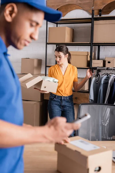 Joven vendedor sosteniendo caja de cartón cerca de la ropa y desenfocado africano americano repartidor en línea tienda web - foto de stock