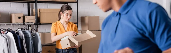 Inhaber hält Schachtel mit Kleidung in der Nähe verschwommener afrikanisch-amerikanischer Zusteller in Online-Webshop, Banner — Stockfoto