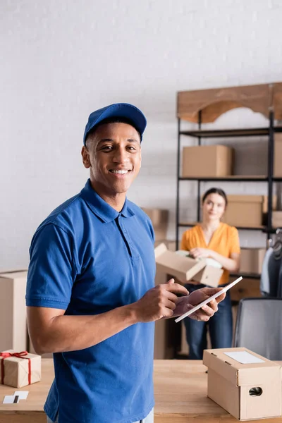 Repartidor afroamericano sonriente sosteniendo tableta digital cerca de cajas en tienda web en línea - foto de stock