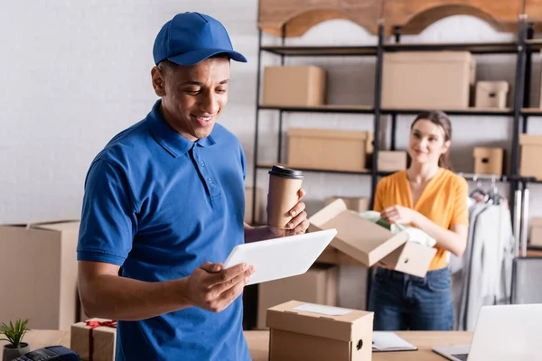 Positive african american deliveryman holding coffee to go and digital tablet near blurred proprietor in online web store — Stock Photo