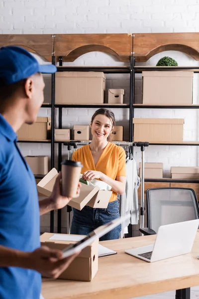 Proprietario sorridente che tiene la scatola con i vestiti vicino al corriere africano americano sfocato con tablet digitale e caffè per andare nel negozio online — Foto stock