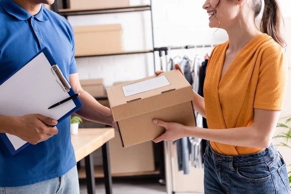 Cropped view of positive proprietor giving carton box to african american deliveryman with clipboard in online web store — Stock Photo