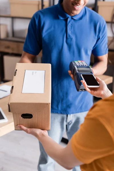 Ausgeschnittene Ansicht des Inhabers, der mit dem Smartphone bezahlt und Karton von einem afrikanischen amerikanischen Kurier im Online-Webshop nimmt — Stockfoto