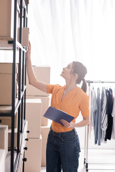 Seitenansicht des Inhabers, der Klemmbrett hält und Karton aus dem Regal im Online-Webshop nimmt — Stockfoto