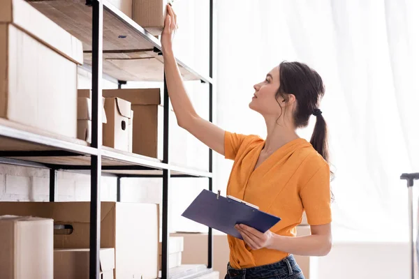 Vista lateral del propietario de la tienda web en línea que sostiene el portapapeles cerca del estante con cajas de cartón - foto de stock