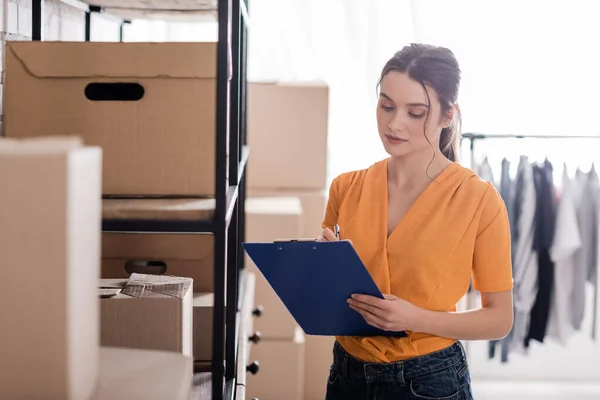 Propietario de la tienda web en línea escribir en portapapeles cerca de cajas de cartón en rack - foto de stock