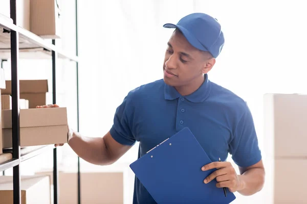 Uomo di consegna afroamericano che tiene appunti e scatola di cartone nel negozio online — Foto stock