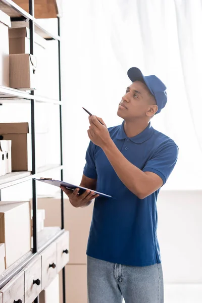 African american courier holding clipboard near packages on rack in online web store — Stock Photo