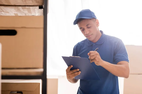 Distribuidor afroamericano escribiendo en portapapeles cerca de cajas de cartón en tienda web en línea - foto de stock