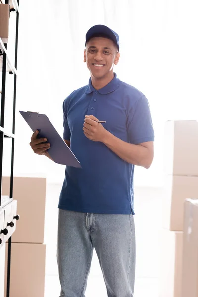 Positive african american deliveryman holding clipboard near cardboard boxes in online web store — Stock Photo