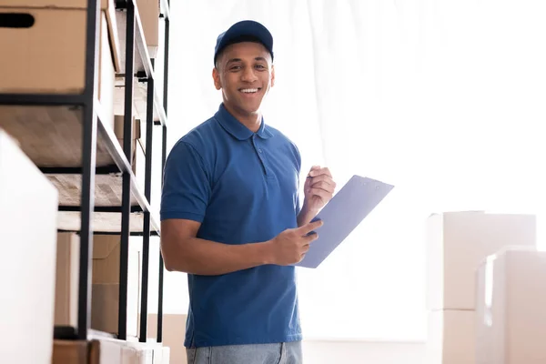 Positive afrikanisch-amerikanische Deliveryman hält Klemmbrett in der Nähe von Rack mit Boxen in Online-Webshop — Stockfoto