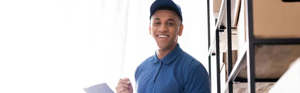 Positive african american deliveryman in uniform holding clipboard and looking at camera near boxes on rack in online web store, banner — Stock Photo