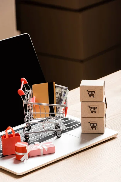 Carrito de compras de juguete con tarjeta de crédito y dinero cerca de paquetes en el ordenador portátil con pantalla en blanco - foto de stock