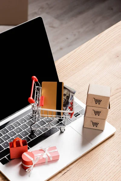 Toy shopping cart with credit card and money near packages on laptop with blank screen — Stock Photo