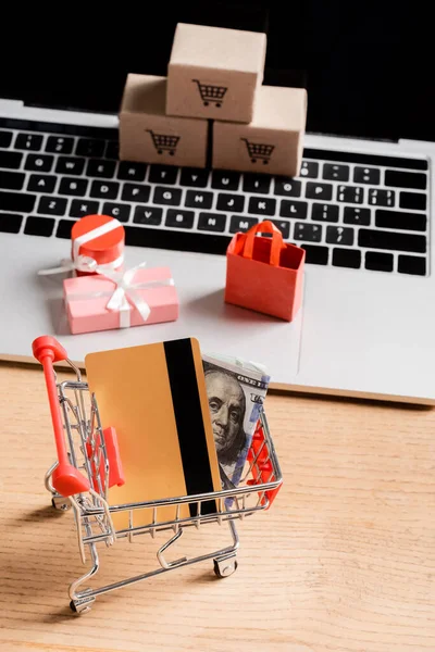 Tarjeta de crédito y dinero en el carrito de compras de juguetes cerca de la computadora portátil con pequeños paquetes en la mesa - foto de stock