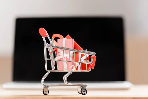 Carrito de compras de juguete con cajas de regalo cerca de la computadora portátil borrosa - foto de stock