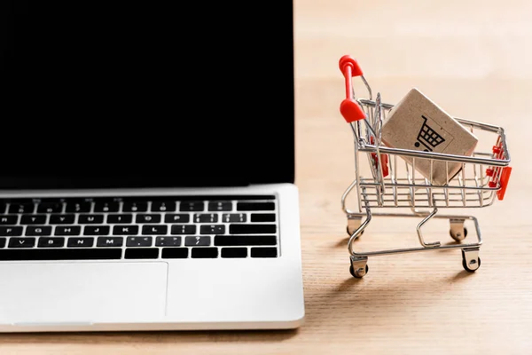 Toy shopping cart with package near laptop with blank screen on table — Stock Photo